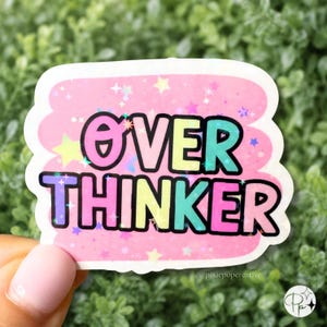 May include: A die-cut sticker featuring the text OVER THINKER in bold, black-outlined letters with a gradient of pink, yellow, and teal. The background is a soft pink with a holographic star pattern and small sparkles throughout the design.