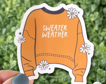 May include: A die-cut sticker featuring an orange long-sleeve sweater with a dark blue collar. The text "SWEATER WEATHER" is printed in white on the chest. The design is decorated with white daisy illustrations and small star accents.