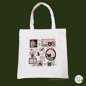 May include: A white tote bag featuring a central graphic print with various illustrations including a stack of books, a ghost reading, a mug labeled Pumpkin Spice, a bat, a witch hat, and the text Book Lover, Just One More Chapter.
