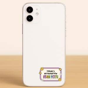 May include: A white smartphone with a sticker that reads "TODAY'S AFFIRMATION STAY PETTY" in a pink, yellow, and white design. The sticker has smiley faces and flowers.