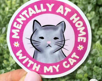 May include: A circular sticker featuring a gray cat illustration centered on a light pink background. A vibrant pink border surrounds the center, containing the white text MENTALLY AT HOME WITH MY CAT and two small white flower icons on the sides.
