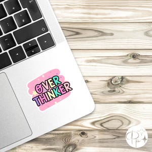 May include: A rectangular sticker applied to the silver surface of a laptop. The sticker features a pink background with small stars and the text OVER THINKER in bold, colorful, bubble-style letters with a black outline and a light blue fill.