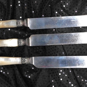 May include: Three butter knives with mother-of-pearl handles and silver blades. The handles have a decorative silver band at the base of the blade. The knives are arranged horizontally against a black background.