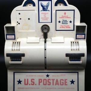 May include: A vintage white metal U.S. Postage stamp vending machine with blue and red text. It features a central handle, two coin return slots, and instructions to deposit three dimes for eight cent stamps, manufactured by American Adjustomatic Corp.