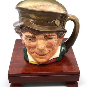 May include: A ceramic mug shaped like a man's head, wearing a brown hat. The mug has a handle and is set on a small, rectangular wooden stand. The man's face is detailed with painted features, including a smile and a green jacket.