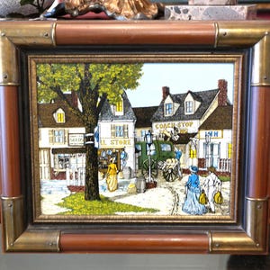 May include: Framed vintage print depicting a quaint town scene with shops, a coach, and figures in period clothing. The frame is brown with brass accents. The print features the words "General Store" and "Coach Stop".