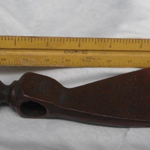 May include: A rusty brown metal tomahawk with a rounded head and a wooden handle. The tomahawk is about 6 inches long and is lying on a white surface.
