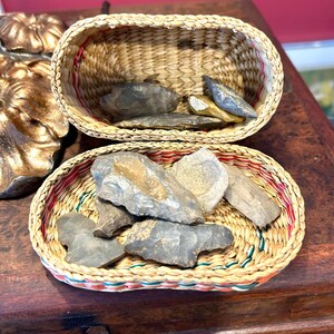 May include: Two woven wicker baskets, each filled with gray and tan stones. The baskets have a natural tan color with red, green, and white trim. The stones vary in size and shape, with some appearing to be chipped or flaked.