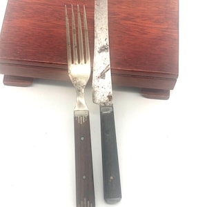 May include: A vintage fork and knife set with dark wooden handles and silver-colored metal blades. The fork has four tines, and the knife has a straight blade. The set is placed on a dark brown wooden surface.