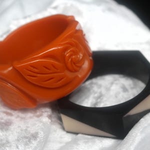 May include: Two bracelets: an orange, carved bangle with a floral design and a black, geometric bracelet with white accents. The orange bracelet features a rose and leaf motif. The black bracelet has a hexagonal shape.