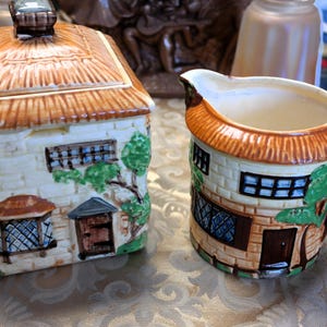 May include: A ceramic sugar bowl and creamer set, each designed to resemble a quaint cottage. The sugar bowl has a thatched roof lid, while the creamer features a handle and spout. Both pieces are painted with brickwork, windows, and doors.