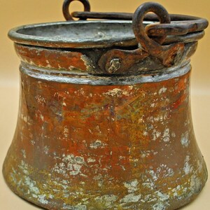 May include: A weathered copper bucket with a flared base and a wide rim. The exterior surface displays a mottled patina with patches of oxidized green, orange, and metallic brown tones. A dark, rusted iron handle is attached to two side loops.