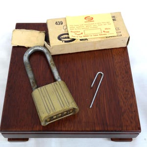 May include: A vintage brass combination padlock with a silver shackle, resting on a dark wooden surface. A small metal tool and a cardboard box with the text "SESAMEE" and "COBB" are also present. The lock has a five-digit combination.