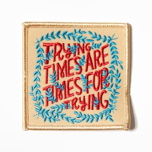 May include: A beige fabric patch with a blue and red embroidered design. The text reads "Trying times are times for trying".
