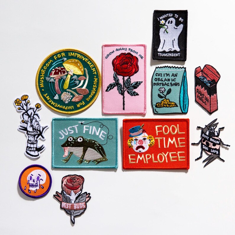Puede incluir: Una colecci&oacute;n de parches bordados con varios dise&ntilde;os y texto. Los dise&ntilde;os incluyen una seta, una rosa, un fantasma, una rana, un payaso y frases como "Just Fine" y "Fool Time Employee". Los parches son de varios colores.