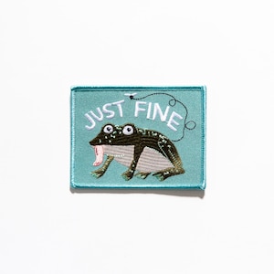 Peut inclure: Un patch brodé bleu clair avec une grenouille et le texte "JUST FINE" en blanc.