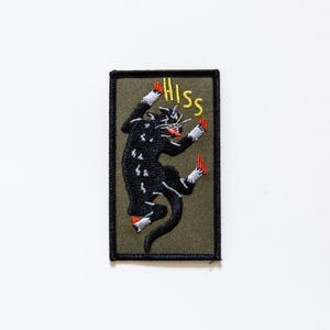 May include: A green embroidered patch with a black cat jumping with its claws out and the word "HISS" in yellow letters.