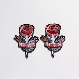 May include: Two embroidered iron-on patches featuring pink roses with green leaves and a banner that reads "BEST BUDS" in white text.