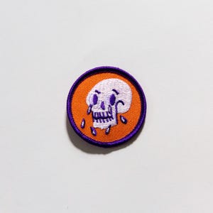 Puede incluir: Parche bordado circular con un dise&ntilde;o de calavera blanca, detalles morados y l&aacute;grimas, sobre un fondo naranja. El parche tiene un borde morado. Adecuado para aplicar en ropa o accesorios.