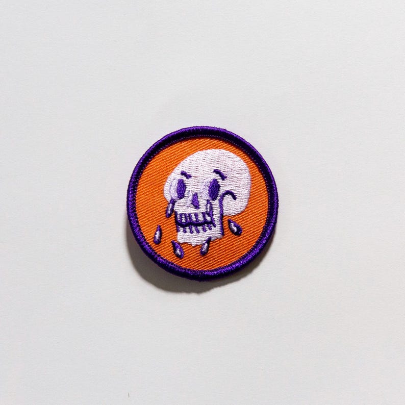 Puede incluir: Parche bordado circular con un dise&ntilde;o de calavera blanca, detalles morados y l&aacute;grimas, sobre un fondo naranja. El parche tiene un borde morado. Adecuado para aplicar en ropa o accesorios.
