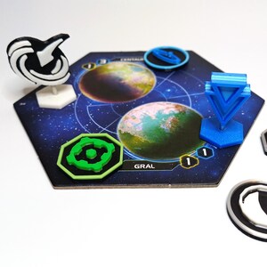 May include: A hexagonal board game tile featuring two planet illustrations, labeled CENTAURI and GRAL. The tile includes a white and black standing marker, a bright blue standing marker, a green octagonal token, and three circular black and silver tokens.