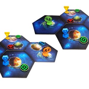 May include: Two clusters of hexagonal board game tiles featuring space-themed illustrations of planets and nebulae. The tiles are arranged in groups, topped with various plastic game pieces in blue, green, yellow, orange, and red colors.