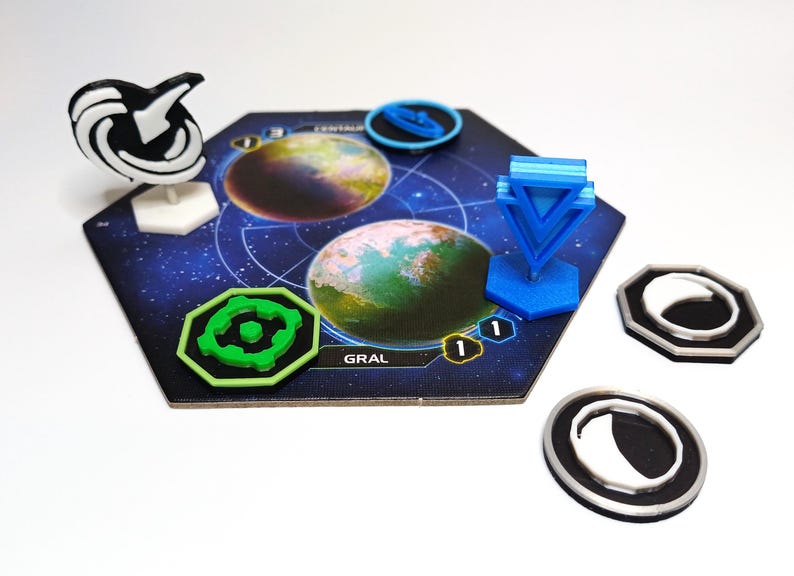 May include: A hexagonal board game tile featuring two planet illustrations, labeled CENTAURI and GRAL. The tile includes a white and black standing marker, a bright blue standing marker, a green octagonal token, and three circular black and silver tokens.