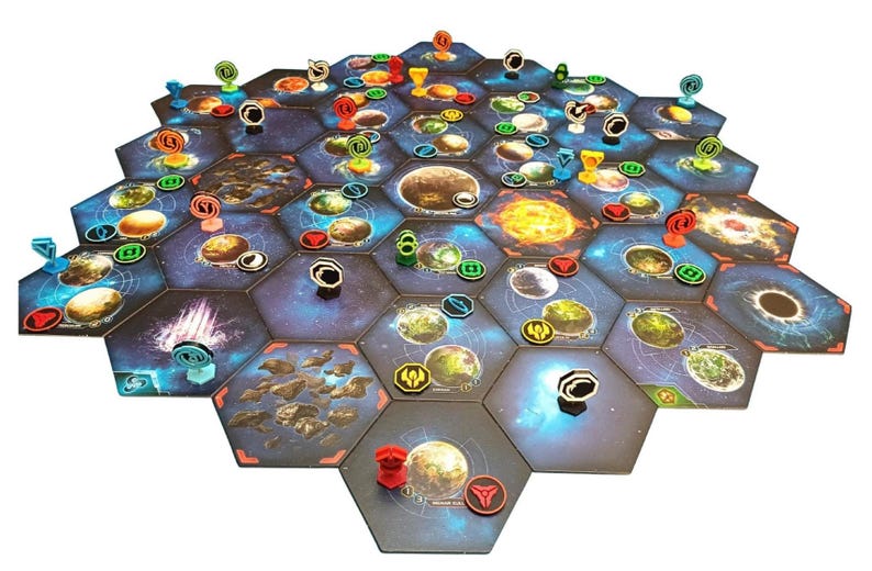 May include: A board game setup featuring a large hexagonal grid of space-themed tiles. The tiles display various planets, stars, and nebulae in shades of blue, orange, and black. Small colored plastic tokens and markers are placed on the tiles.