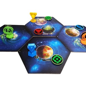 May include: A board game setup featuring three hexagonal space-themed tiles with dark blue galaxy backgrounds. The tiles display planetary illustrations, including one labeled Thibah. Various colored plastic game pieces and tokens are placed on top.