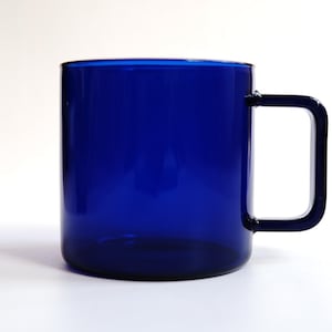 May include: A translucent cobalt blue glass mug featuring a cylindrical body and a distinct square-shaped handle. The vessel is constructed from smooth, solid-colored glass, showcasing a deep blue hue throughout the entire structure of the cup.