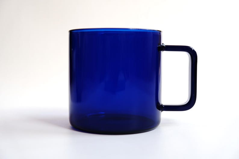 May include: A translucent cobalt blue glass mug featuring a cylindrical body and a distinct square-shaped handle. The vessel is constructed from smooth, solid-colored glass, showcasing a deep blue hue throughout the entire structure of the cup.