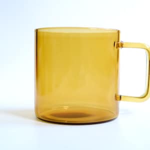 May include: A translucent amber-colored glass mug with a cylindrical body and a square-shaped handle. The mug features a smooth, polished finish and a consistent golden-brown hue throughout the glass, showcasing a simple and functional design.