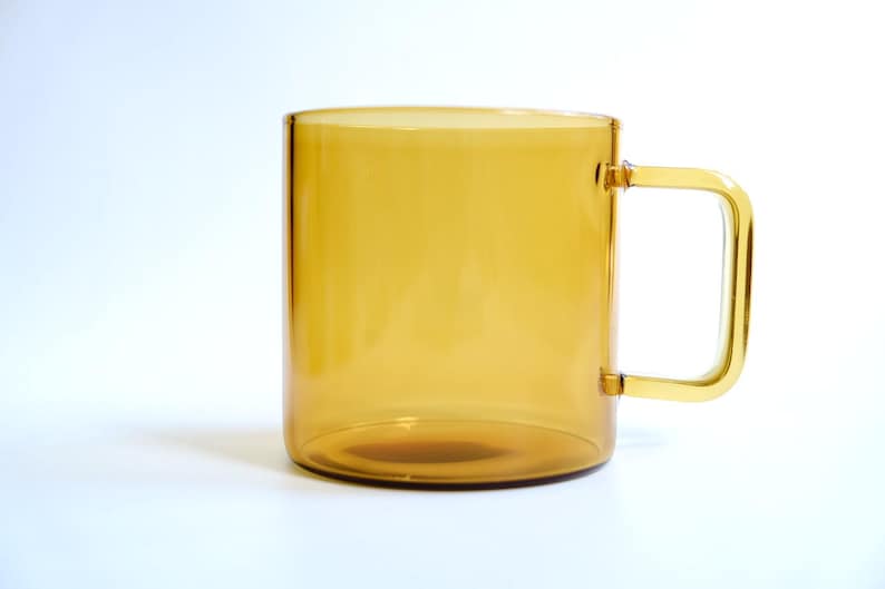May include: A translucent amber-colored glass mug with a cylindrical body and a square-shaped handle. The mug features a smooth, polished finish and a consistent golden-brown hue throughout the glass, showcasing a simple and functional design.