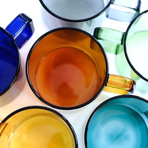 May include: A collection of six translucent glass mugs arranged in a circular pattern, viewed from directly above. The mugs feature vibrant colors including deep blue, amber, light green, yellow, teal, and clear glass, each with a matching handle.
