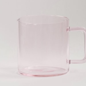 May include: A translucent light pink glass mug with a cylindrical body and a square-shaped handle. The mug features a smooth, clear finish and a simple, functional design with a flat base and a straight rim, showcasing a uniform pale pink hue.