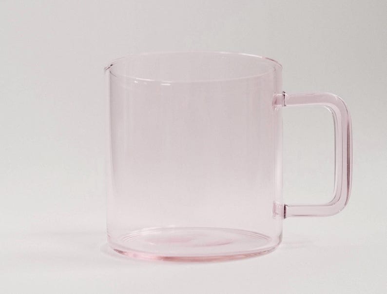May include: A translucent light pink glass mug with a cylindrical body and a square-shaped handle. The mug features a smooth, clear finish and a simple, functional design with a flat base and a straight rim, showcasing a uniform pale pink hue.