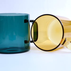 May include: Two translucent glass mugs with simple cylindrical bodies and flat handles. One mug is a deep teal blue color, standing upright, while the second mug is a warm amber yellow color, positioned on its side to show the interior.