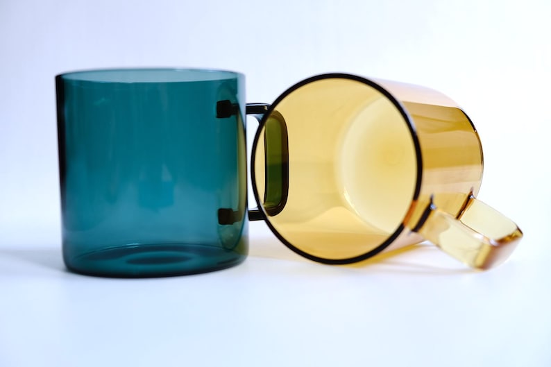May include: Two translucent glass mugs with simple cylindrical bodies and flat handles. One mug is a deep teal blue color, standing upright, while the second mug is a warm amber yellow color, positioned on its side to show the interior.