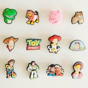 May include: A selection of vibrant cartoon character charms, featuring a green dinosaur, a dog, a pink pig, and a brown horse. Other charms include Woody, Jessie, Buzz Lightyear, and the Toy Story logo. These charms are likely for shoes or accessories.