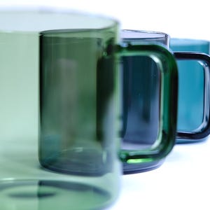 May include: A row of four translucent glass mugs arranged in a line, each featuring a distinct shade of green. The mugs have a cylindrical body and a square-shaped handle. The colors transition from a light sage green to a deep, dark forest green.