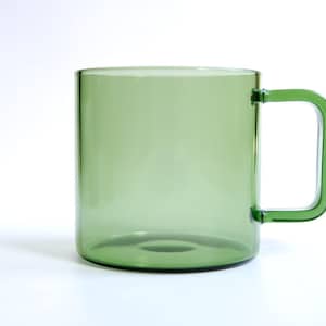 May include: A translucent sage green glass mug with a cylindrical body and a square-shaped handle. The mug features a smooth, polished finish and a clear, uniform color throughout the glass, showcasing a simple and functional design for beverages.