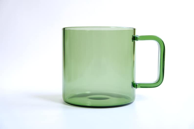 May include: A translucent sage green glass mug with a cylindrical body and a square-shaped handle. The mug features a smooth, polished finish and a clear, uniform color throughout the glass, showcasing a simple and functional design for beverages.
