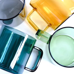 May include: A collection of four translucent glass mugs in shades of dark gray, amber, deep teal, and light green. Each mug features a cylindrical body and a square-shaped handle, arranged in a top-down view to showcase the vibrant colors.