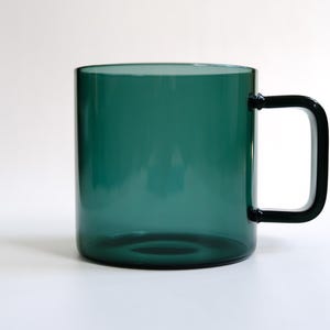 May include: A translucent dark green glass mug with a cylindrical body and a matching dark green rectangular handle. The mug features a smooth, polished finish and a simple, clean design, showcasing the deep emerald hue of the glass material.