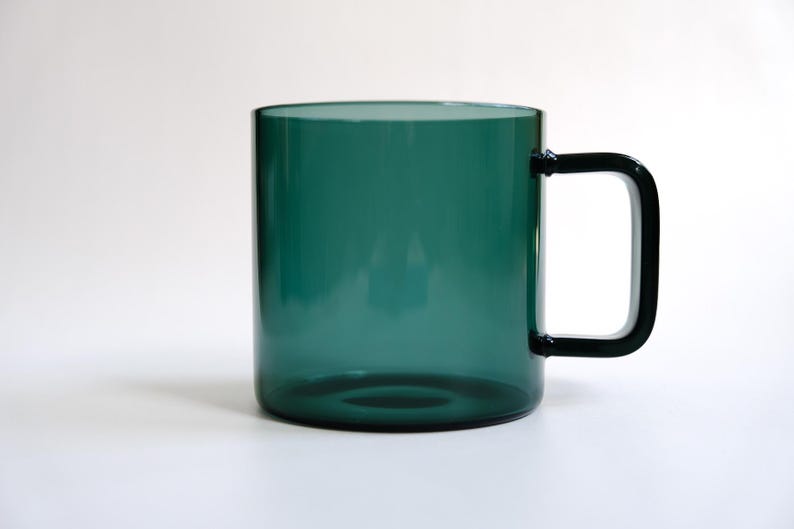 May include: A translucent dark green glass mug with a cylindrical body and a matching dark green rectangular handle. The mug features a smooth, polished finish and a simple, clean design, showcasing the deep emerald hue of the glass material.
