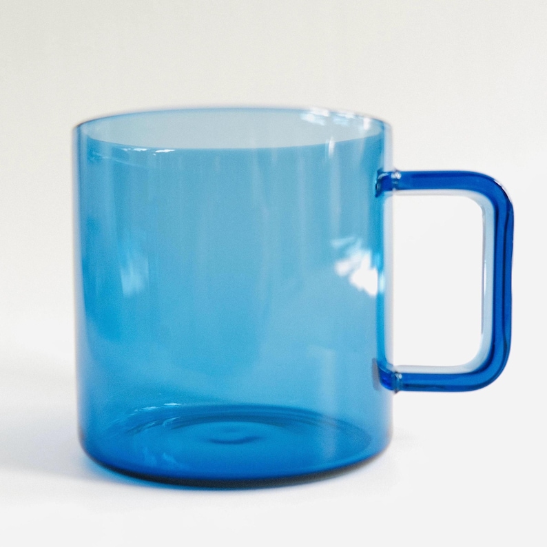 May include: A translucent blue glass mug with a cylindrical body and a square-shaped handle. The mug features a smooth, clear finish that allows light to pass through, highlighting the vibrant blue color of the glass material throughout the vessel.