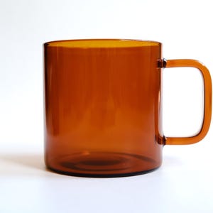 May include: A translucent amber-colored glass mug with a cylindrical body and a square-shaped handle. The mug features a smooth, polished finish and a simple, clean design, showcasing the warm, brownish-orange hue of the glass material.