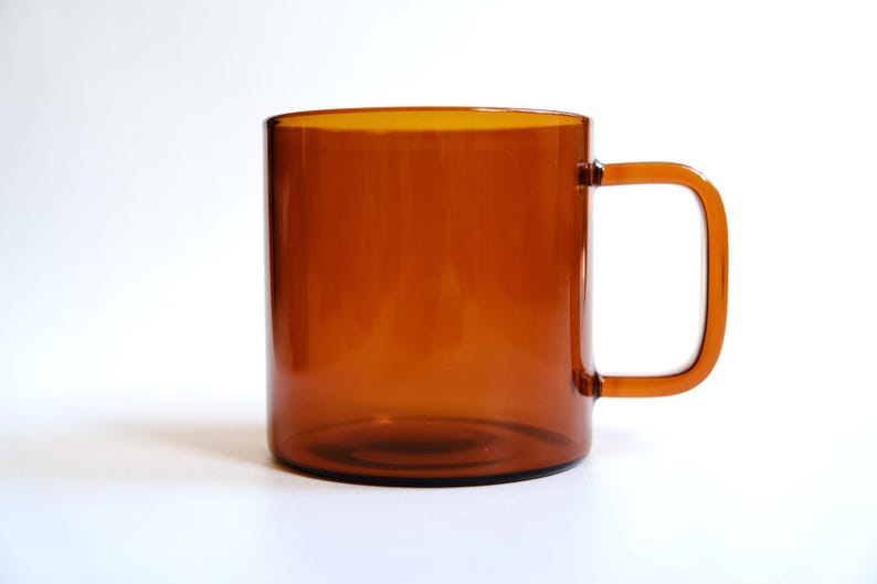 May include: A translucent amber-colored glass mug with a cylindrical body and a square-shaped handle. The mug features a smooth, polished finish and a simple, clean design, showcasing the warm, brownish-orange hue of the glass material.