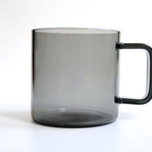 May include: A translucent, smoke-gray glass mug featuring a cylindrical body and a distinct, angular, square-shaped handle attached to the side. The glass has a smooth, uniform finish, allowing light to pass through the dark, neutral-toned material.