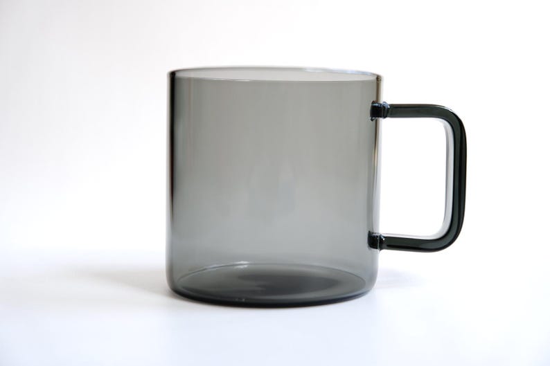 May include: A translucent, smoke-gray glass mug featuring a cylindrical body and a distinct, angular, square-shaped handle attached to the side. The glass has a smooth, uniform finish, allowing light to pass through the dark, neutral-toned material.